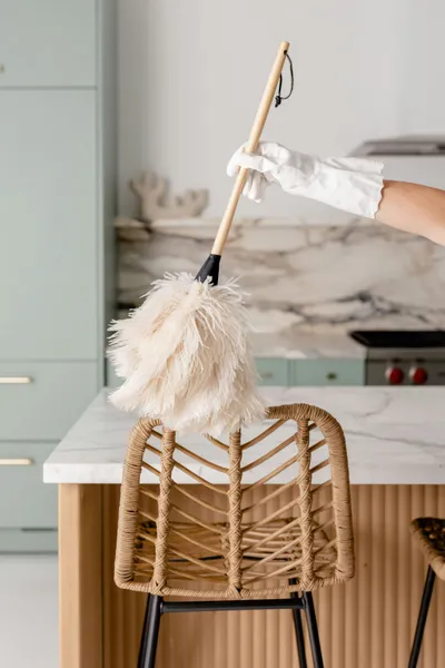 Main gantée passant un plumeau en plumes sur une chaise en rotin, dans une cuisine claire : ménage à domicile